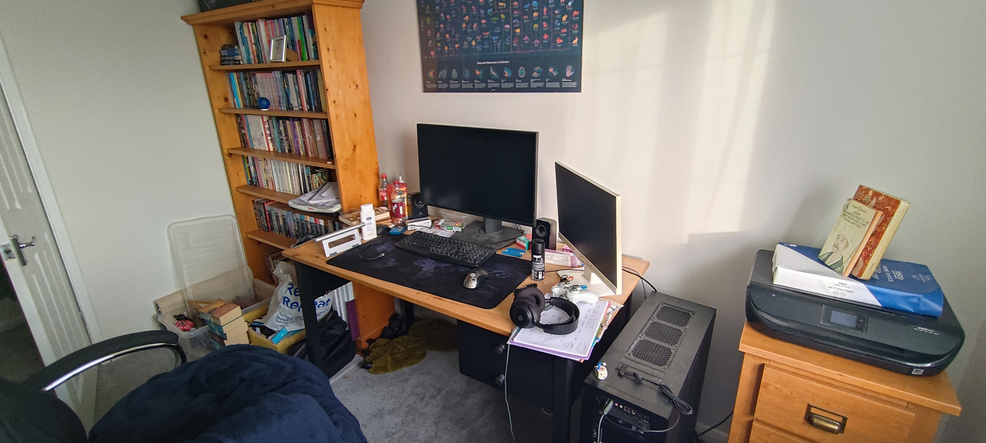 the full study a view of our full, cluttered study; a bookshelf, large desk with two monitors, huge desktop PC, and small set of drawers with a printer sat on top, all along one wall with a large office chair partially in frame. The desk is covered in small clutter such as over-the-counter medicine boxes, paperwork, peripherals, and evidence of snacks and drinks.